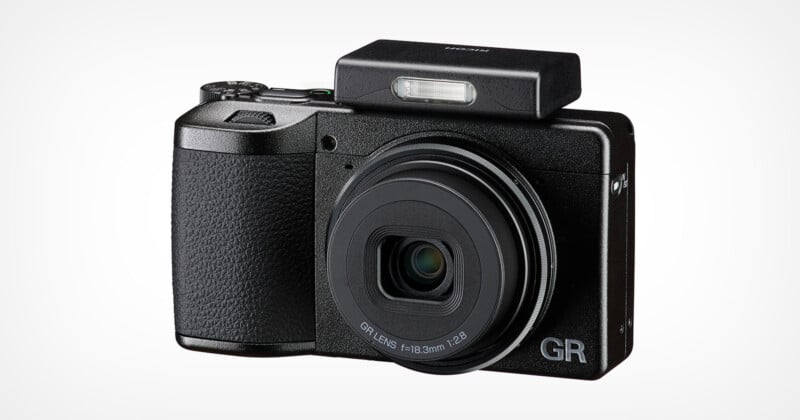 A compact black digital camera with a textured grip, extended lens, and raised rectangular pop-up flash. The letters "GR" are visible on the lower right corner of the camera front.