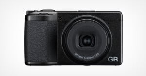 A black compact digital camera with a textured grip, large circular lens labeled "GR LENS f=18.3mm 1:2.8", and the letters "GR" in silver on the bottom right corner of the front.