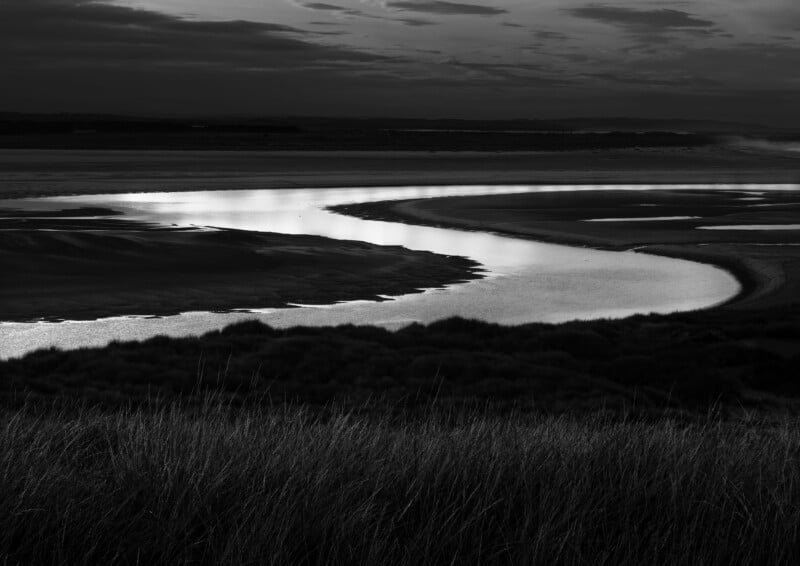 Un río sinuoso serpentea a través de un paisaje oscuro bajo un cielo nublado, reflejando la luz, con hierba alta en primer plano y tierra lejana apenas visible en el horizonte.