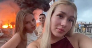 Three young people take a selfie in front of a fire with large black smoke rising in the background, appearing relaxed despite the emergency behind them.
