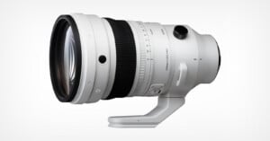 A large, white telephoto camera lens with a tripod mount and various control switches, displayed against a plain white background.