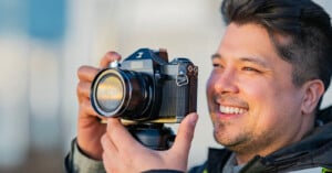 A man smiles while holding a vintage film camera up to his face, ready to take a photo outdoors in natural light.