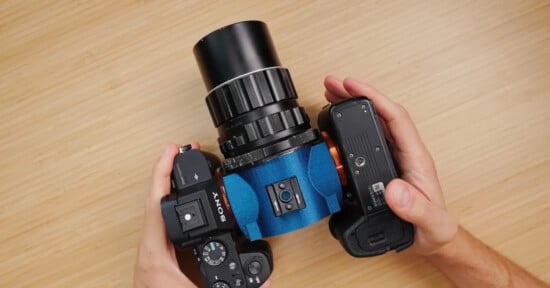 A person holds a Sony camera attached to a large, blue, cylindrical lens with black and silver rings over a light wood surface.