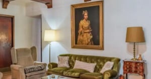 A vintage living room with a green velvet sofa, cushioned armchair, wooden side table, two lamps, and a large framed portrait of a woman in a gold dress hanging on a white wall.