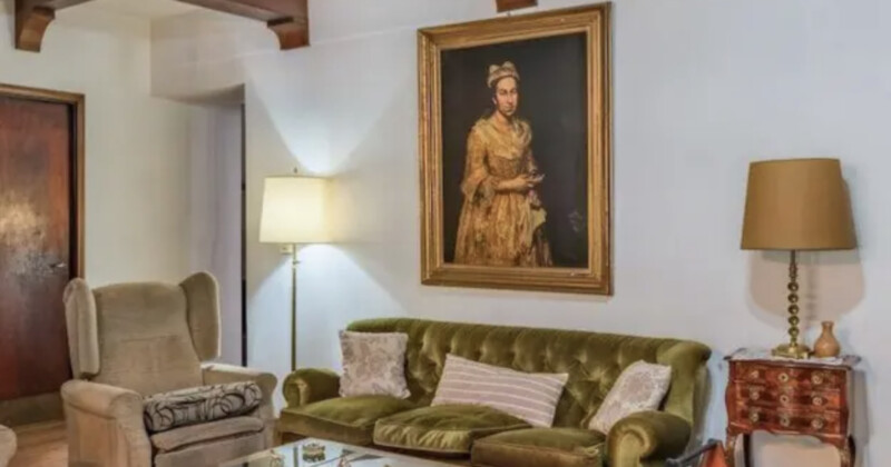 Un gran retrato de una mujer con un vestido dorado colgando de una pared blanca con un sofá de terciopelo verde, sillón acolchado, mesa de madera, dos lámparas y un gran retrato enmarcado de una mujer con un vestido dorado colgando de una pared blanca.