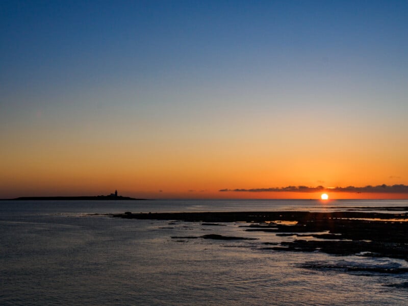 Sunset over a calm sea with an orange sky. The sun is low on the horizon, and a small island with a lighthouse is visible in the distance to the left. The foreground shows dark, rocky tidal pools.