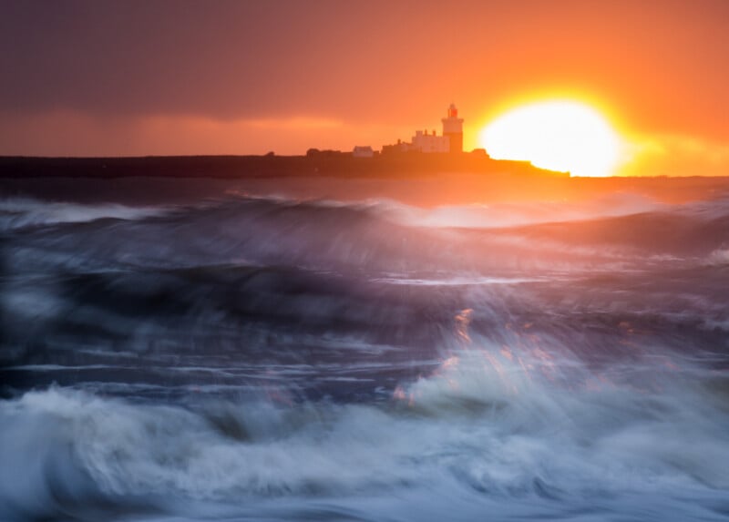 A dramatic sunset illuminates a lighthouse and distant buildings on a dark coastline, with large, blurred ocean waves crashing in the foreground under a vibrant orange sky.