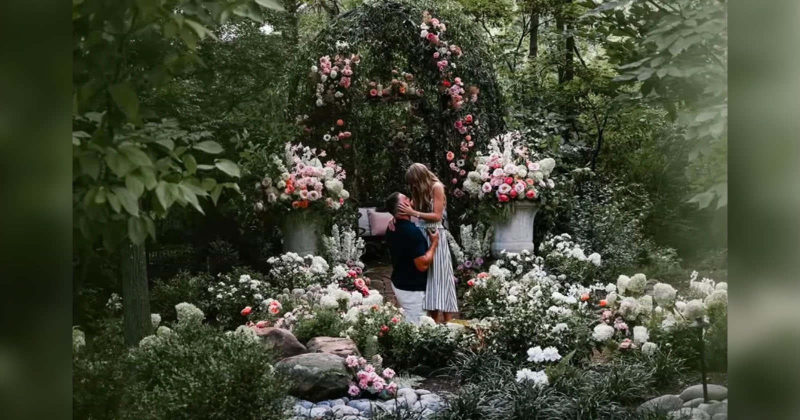A woman in a striped dress holds a child in a lush garden filled with blooming flowers and greenery; large floral arrangements and an arch create a romantic, whimsical atmosphere.