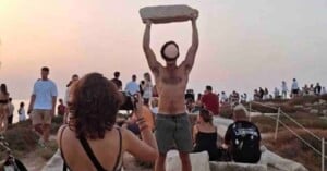 A shirtless man stands outdoors, holding a large stone slab above his head as a crowd of people gather around. A woman in the foreground takes a photo of him. It's an open area with rocks and a rope fence.