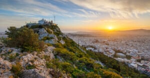 White chapel atop Lycabettus Hill overlooks Athens at sunset, with rocky terrain and green foliage in the foreground and city buildings sprawling to the horizon under a colorful sky.