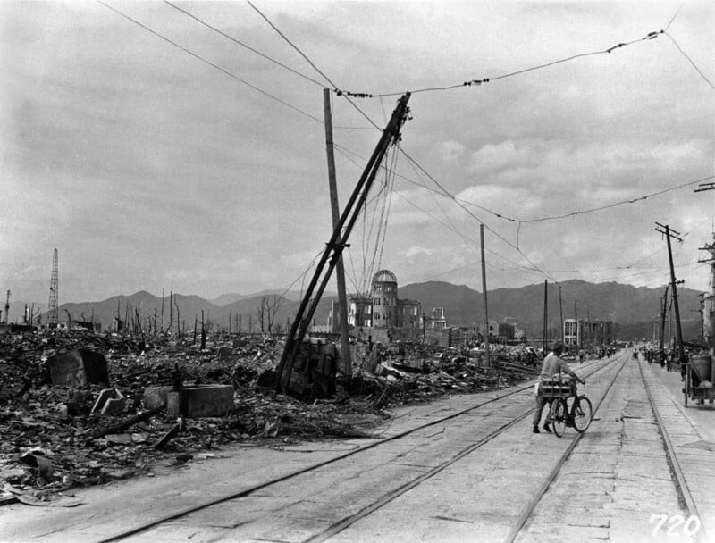 A man walks his bicycle down a deserted street lined with debris and ruined buildings in Hiroshima after the atomic bombing; damaged power lines lean over the road under a cloudy sky.