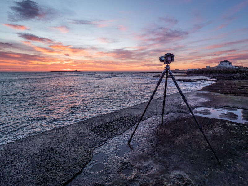 La cámara del trípode se erigió en la superficie húmeda de concreto del mar al atardecer, capturando cielos coloridos y olas tranquilas. Los edificios son visibles en el fondo de la costa.