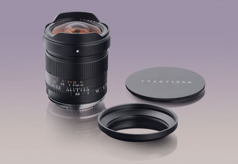 A TTArtisan camera lens stands upright on a reflective surface, with its front cap and lens hood placed beside it against a soft gradient background.