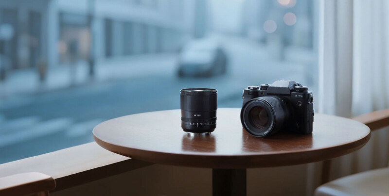 A camera and a separate lens sit on a round wooden table by a window, with a blurred city street and buildings visible outside in the background.