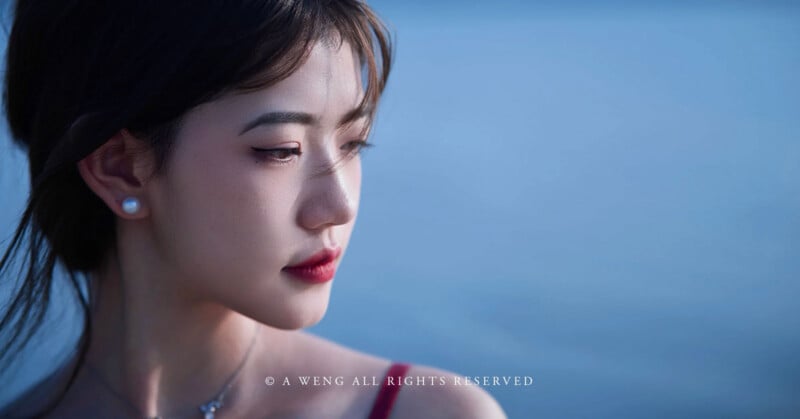 A young woman with long dark hair and pearl earrings gazes downward against a soft blue background, with gentle lighting highlighting her face.