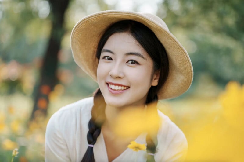A young woman with braided hair and a straw hat smiles warmly while surrounded by yellow flowers in an outdoor garden. The background is green and softly blurred.