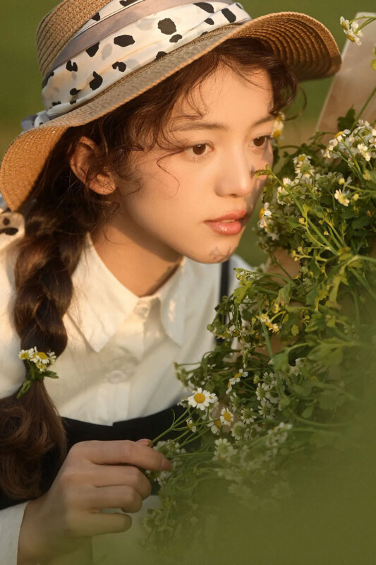 A young woman with wavy hair in a braid, wearing a straw hat with a patterned ribbon and a white blouse, gently holds wildflowers while gazing thoughtfully at a bouquet of small white daisies in a sunlit outdoor setting.