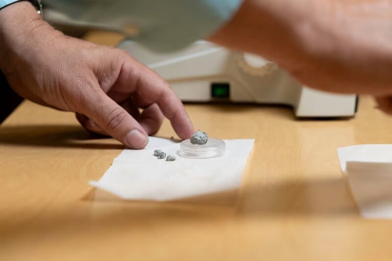 A person’s hands examine small gray rock samples on a petri dish placed on white paper atop a wooden table, with lab equipment visible in the background.