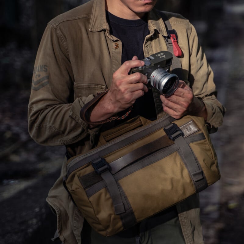 A person wearing a brown jacket and crossbody bag stands outdoors, holding and adjusting a compact camera in their hands. The scene appears to be in a natural or urban setting with subdued lighting.
