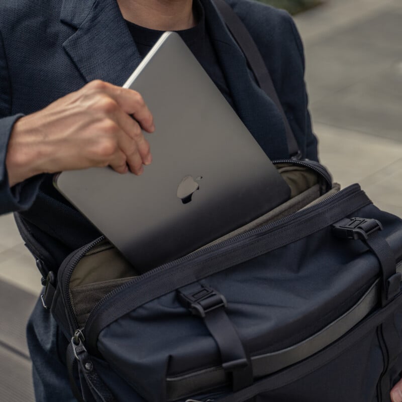 A person wearing a dark blazer places a silver laptop into a black shoulder bag outdoors.
