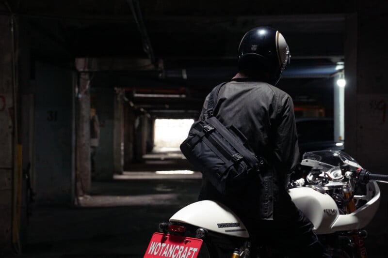 A person wearing a helmet and dark jacket sits on a white motorcycle with a red license plate in a dimly lit parking garage, carrying a black sling bag over their shoulder.