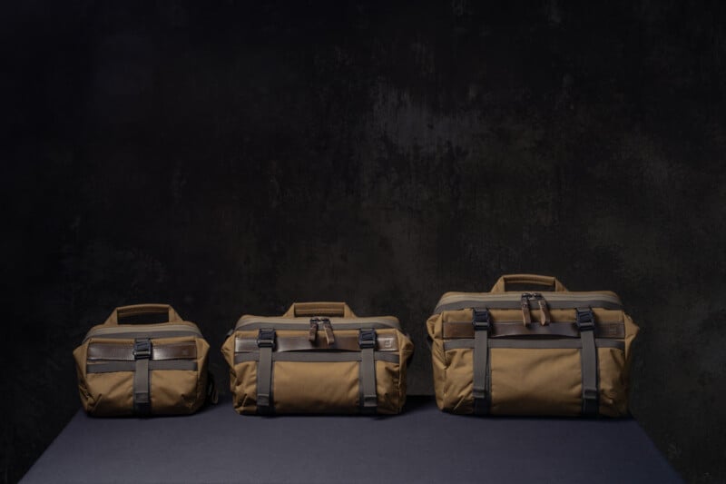 Three brown travel bags of different sizes are lined up on a dark surface against a black textured background, each featuring handles, pockets, and black buckle straps.