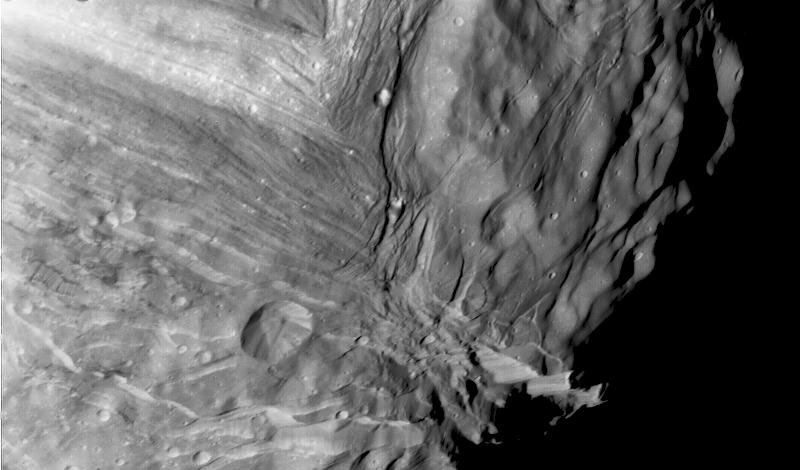Close-up image of a heavily cratered, grooved, and ridged surface of a celestial body, possibly a moon or asteroid, with deep shadows and varying textures visible.