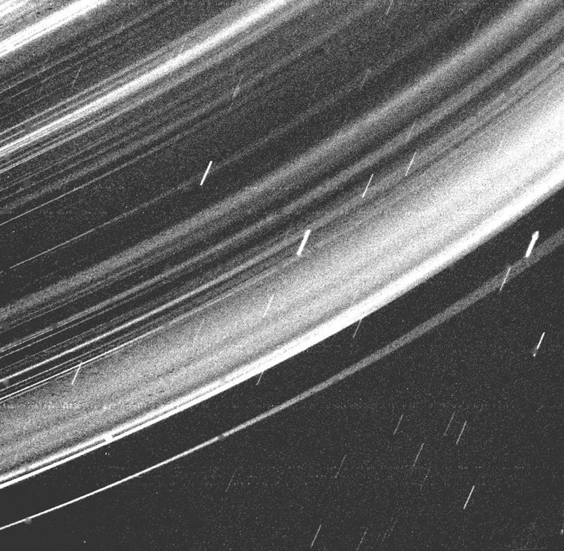 Black and white image showing the rings of Saturn with bright, curving lines set against a dark background, and several smaller streaks moving diagonally, representing moons or particles.