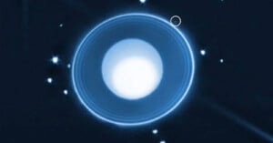A bright, white, circular planet with thin blue rings is surrounded by small white stars on a dark background. A faint, small dot near the upper right ring is highlighted with a circle.