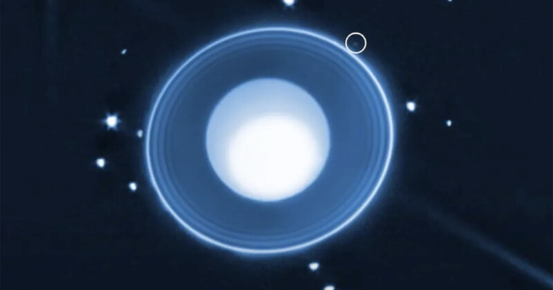A bright, white, circular planet with thin blue rings is surrounded by small white stars on a dark background. A faint, small dot near the upper right ring is highlighted with a circle.