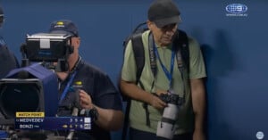 Two photographers stand courtside at a tennis match. One operates a large TV camera, while the other holds a professional camera with a telephoto lens. A scoreboard shows Medvedev vs. Bonzi with Bonzi at match point.
