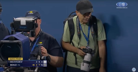 Two photographers stand courtside at a tennis match. One operates a large TV camera, while the other holds a professional camera with a telephoto lens. A scoreboard shows Medvedev vs. Bonzi with Bonzi at match point.