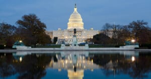 The U.S. Capitol building illuminated at dusk is reflected in a calm pool of water, surrounded by trees and statues in Washington, D.C.