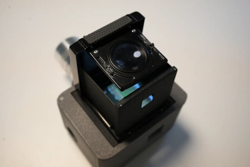 A close-up of a compact black optical device with a lens on top and a small rectangular opening on the side, placed on a light-colored surface.
