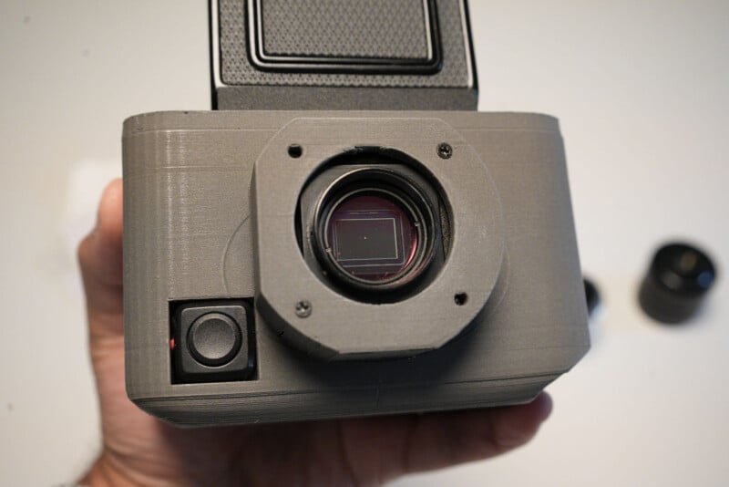 A close-up of a person holding a gray, boxy camera with an exposed sensor and no lens attached. The background is neutral, with two blurred lenses visible to the side.