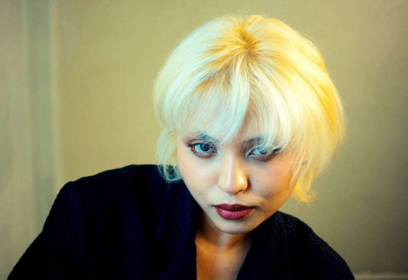 A person with short, platinum blonde hair and bangs looks intently at the camera. They are wearing a dark textured jacket and have bold eye makeup and dark red lipstick, set against a neutral background.