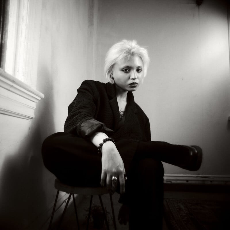 A person with short, light blond hair sits on a chair in a dimly lit room, wearing dark clothing. They look directly at the camera with a serious expression, one leg crossed over the other.