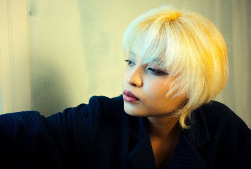 A person with short, bleached blonde hair and wearing a textured dark jacket looks thoughtfully out of frame against a softly lit, neutral background.