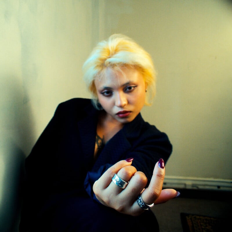 A person with short, bleached blonde hair and dark clothing sits indoors, extending their hand forward with rings on their fingers, giving a confident, intense look toward the camera.