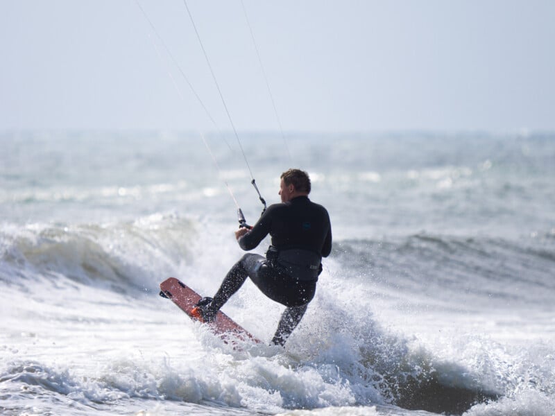 Un hombre lleva un tablero de cometas de traje de buceo en el océano, montando la barra de control mientras monta la barra de control, el agua salpica y el océano se extiende hacia el fondo.