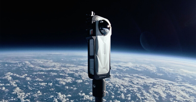 A sleek, modern camera or observation device floats above Earth in space, with the planet’s curved horizon, clouds, and deep blue atmosphere visible below against the darkness of space.