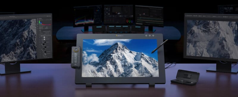 A digital artist’s workspace with three monitors displays a snowy mountain image; the central display is a drawing tablet with a stylus and remote, with editing software visible on the side monitors.