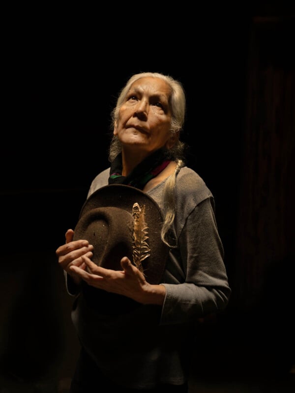 An elderly woman with long gray hair holds a hat with a feather, looking upward thoughtfully. She wears a gray top and a scarf, highlighted by soft, dramatic lighting against a dark background.