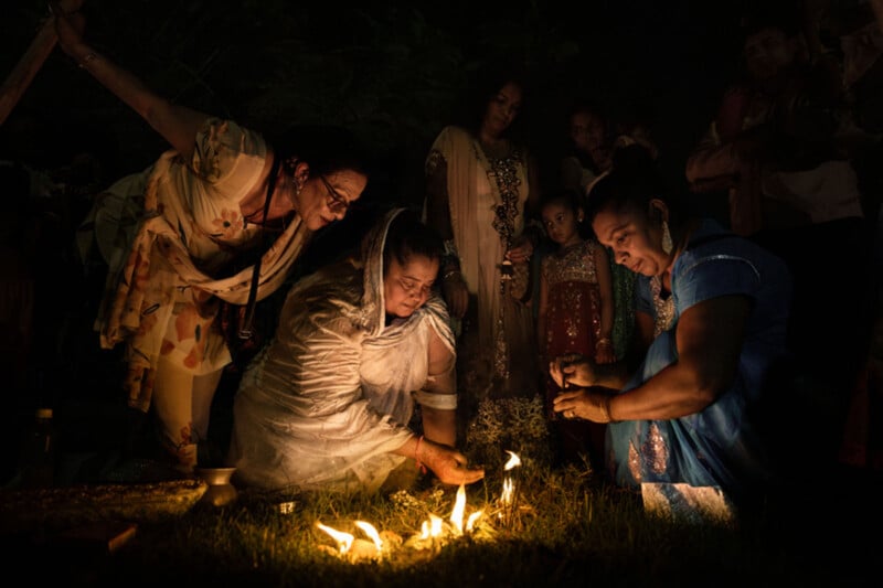 A group of women, dressed in colorful traditional clothing, kneel on the grass at night, lighting candles together in a solemn outdoor ceremony, while others watch in the background.