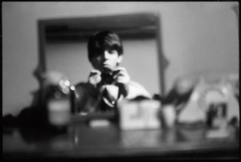 A blurry black-and-white photo shows a person taking a picture of themselves in a mirror, with various indistinct objects on a dresser in the foreground.