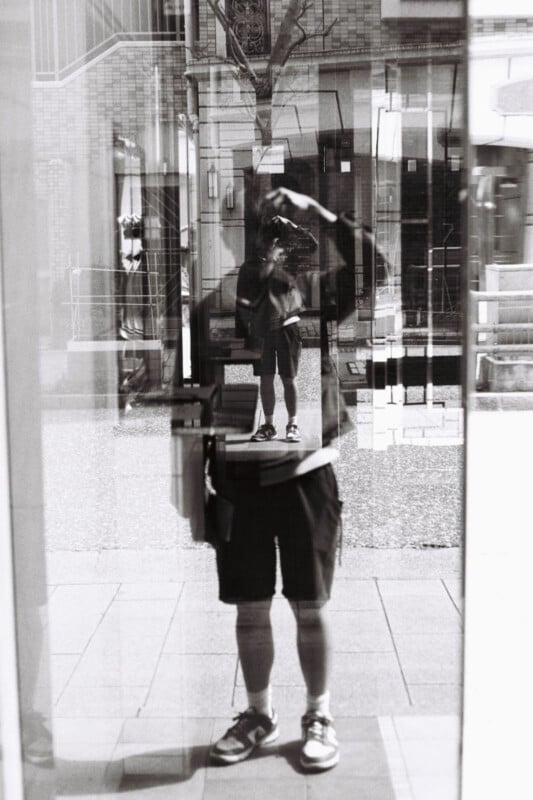 A person wearing shorts and sneakers is taking a photo of their reflection in a glass window, creating layered, overlapping reflections on a city sidewalk. The image is in black and white.