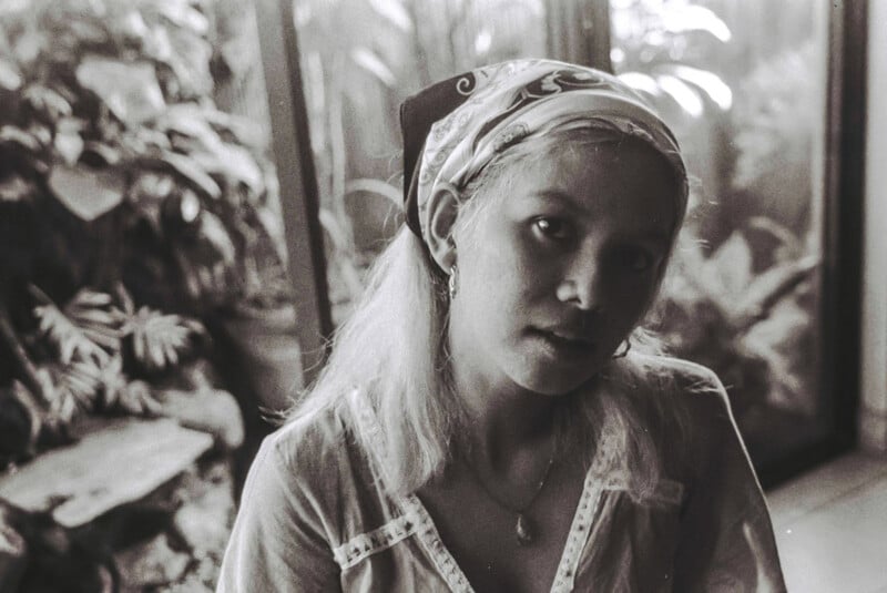 Black and white photo of a woman with light hair wearing a bandana and earrings, sitting indoors near large windows with plants visible outside. She looks slightly away from the camera in soft, natural light.