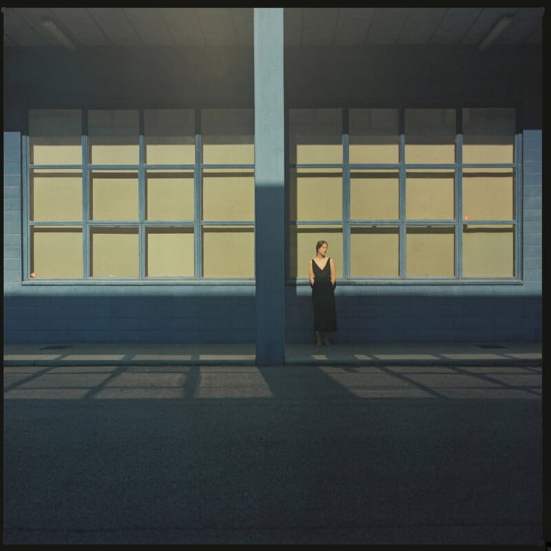 A woman in a black dress stands alone against a blue building with large square windows, partially shaded by the structure’s overhead beams, creating geometric shadows on the ground.