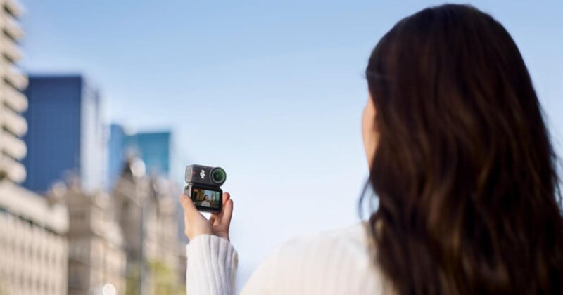 A person with long brown hair holds up a compact camera, filming a cityscape with modern buildings under a clear blue sky. The camera’s screen shows a preview of the scene being recorded.