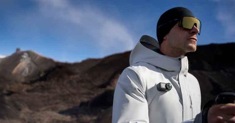 A person wearing a white outdoor jacket, black beanie, and reflective sunglasses stands in a rocky, mountainous landscape under a clear blue sky.